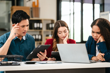 Business team meeting with stressed man rubbing his forehead, while two women review documents, appearing serious and focused.