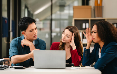Business team meeting with stressed man rubbing his forehead, while two women review documents, appearing serious and focused.