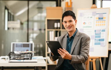 Attractive professional Asian businessman with tablet in office environment, confidently smiling, possibly analyzing data as Capital Market Analyst.