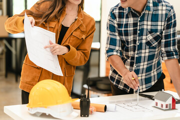 Asian man and Caucasian woman, both engineers, examining set of blueprints closely during team meeting, with house model on the table.
