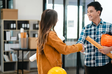 Asian man and Caucasian woman, both engineers, examining set of blueprints closely during team meeting, with house model on the table. Shaking hands