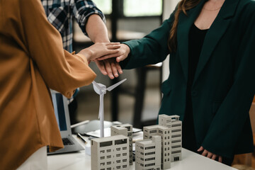 three people place their hands together above a model of buildings with a white wind turbine. teamwork, join hands