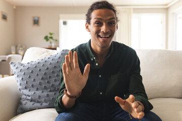 Happy biracial man with long curly hair having video call and smiling in sunny living room at home