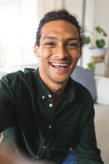 Happy biracial man with long curly hair having video call and laughing in sunny living room at home