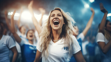 A female sports fan is happy with a group of friends, many cheering together happily and excited to watch their favorite football team. Cheering sports fans wear blue and white cheer team shirts.
