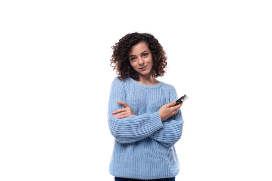 authentic smiling young brunette woman with curls dressed in a blue knitted sweater holds a telephone in her hands