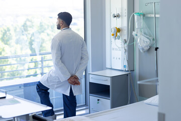 Back of biracial male doctor looking out window in sunny hospital room