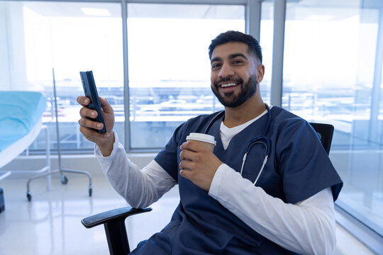 Portrait of happy biracial male doctor using smartphone and holding cup of coffee in hospital office