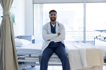 Portrait of happy biracial male doctor sitting on bed in sunny hospital room