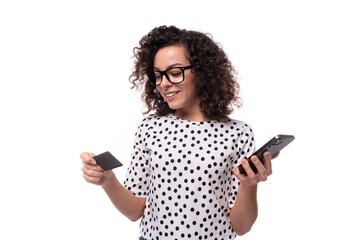 young well-groomed slender lady with black curly hair in a polka-dot blouse holds a plastic card and a smartphone