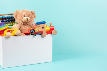 Toy box full of baby kid toys. Container with teddy bear, fluffy and educational wooden toys on light blue background. Cute toys collection for small children. Front view - obrazy, fototapety, plakaty