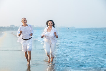 Happy senior indian couple wearing white cloths running together at beach and having fun. Vacations...