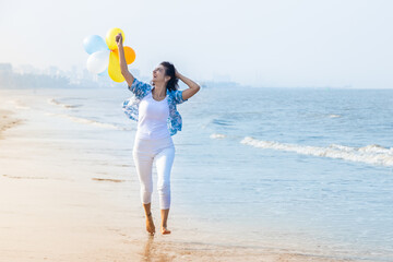 Beautiful young indian woman running with multicolor balloon in hands at the beach. Young girl having fun, holidays and vacation. - obrazy, fototapety, plakaty