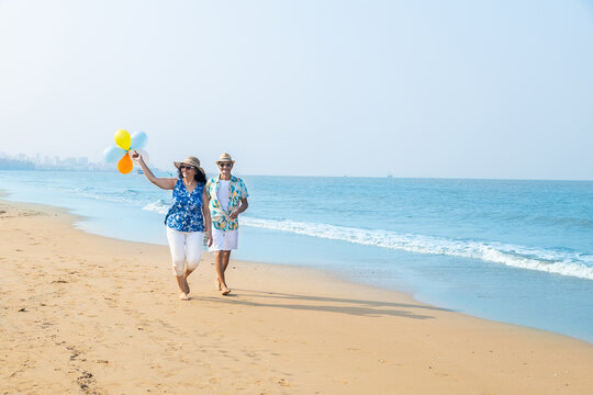 Senior indian couple with colorful balloon in hands running together at the beach. Enjoying vacation, holiday at beach. Copy space. Retirement life.