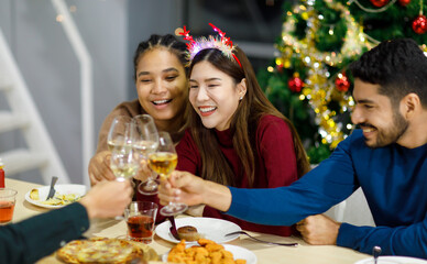 Group of Asian Indian happy cheerful male female friends wears reindeer antlers headband cheers toasting champagne beverage glasses together on dinner table celebrating Christmas eve in dining room
