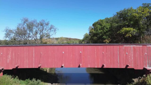 Hogback covered bridge in Madison County Iowa. Low angle aerial shot, from below the side of the bridge, rises over the top and follows the stream. Bright sunny afternoon.