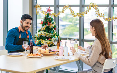Indian male boyfriend Asian female girlfriend lover couple eating delicious tasty fast foods donut cupcake pizza together in dining room full decorated with Xmas tree celebrated Christmas Eve festive