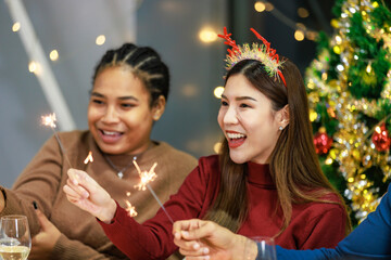 Asian Indian male female friends wears reindeer antlers headband sitting holding playing small sparkler firework celebrating together at dinner table full of food snack in dining room with Xmas tree