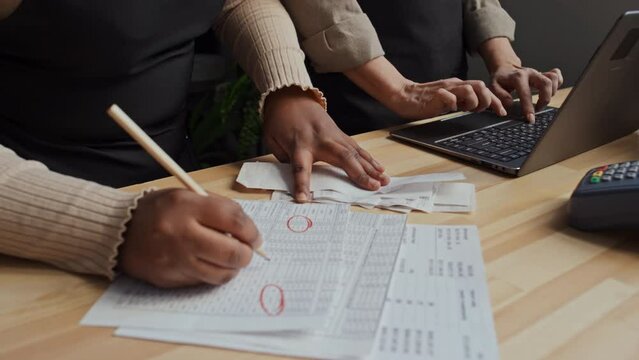 Two small business owners working behind counter while reconciling their accounts using receipts, cheques and laptop
