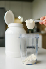 Young man with measuring spoon puts portion of whey protein powder into a shaker on wooden table,...