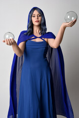 Close up portrait of beautiful female model wearing elegant fantasy blue ball gown, flowing cape with hooded cloak.
 Holding a crystal glass seer orb. Isolated on white studio background.