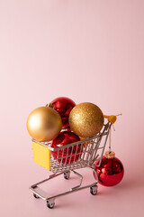 Christmas balls, toys in mini trolley from supermarket on pink background
