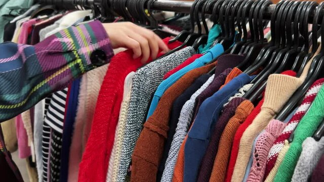 A young buys clothes in a second-hand shop. Thrift store hangers with colorful sweaters. The concept of buying used clothes, fast fashion industry waste reduce and sustainability. Close-up.
