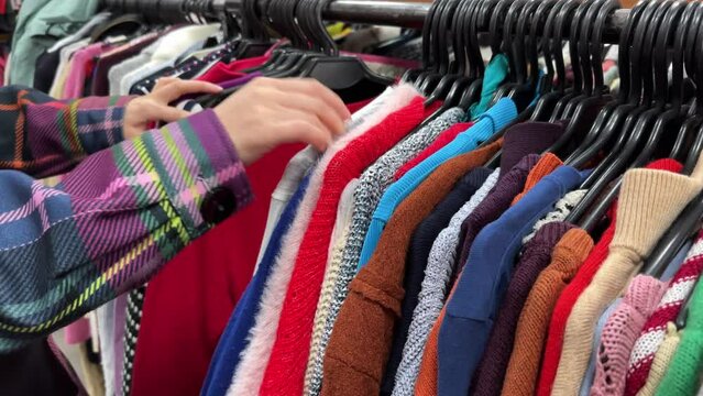 A young woman chooses clothes in a second-hand shop. Thrift store hangers with colorful sweaters. The concept of buying used clothes, fast fashion industry waste reduce and sustainability. Close-up.