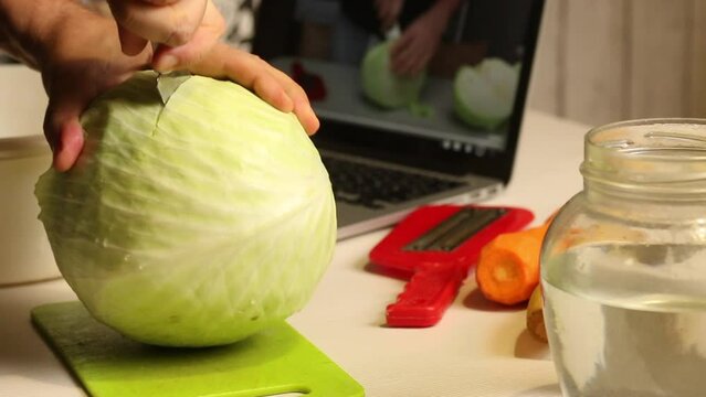 A man watches a video with a step -by -step recipe on a laptop. Preparing sauerkraut at home. He cuts cabbage, removes the stump. Close-up.
