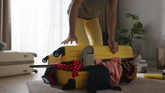 In flurry, woman attempts to close her yellow suitcase, filled with jumble of items. Haste and carelessness lead to messy packing situation. Randomness prevails. Packing bags for journey