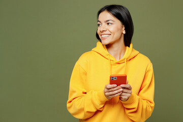 Young smiling happy Latin woman she wear yellow hoody casual clothes hold in hand use mobile cell phone look aside on area isolated on plain pastel green background studio portrait. Lifestyle concept