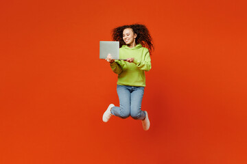 Full body young fun IT woman of African American ethnicity she wear green hoody casual clothes jump high hold use work on laptop pc computer isolated on plain red orange background. Lifestyle concept.