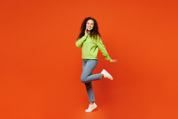 Full body side view nice happy young woman of African American ethnicity she wear green hoody casual clothes put hand on face raise up leg isolated on plain red orange background. Lifestyle concept.