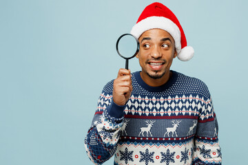 Young smiling man wear knitted sweater Santa hat posing hold in hand use magnifying reading glass isolated on plain pastel blue background. Happy New Year 2024 celebration Christmas holiday concept.