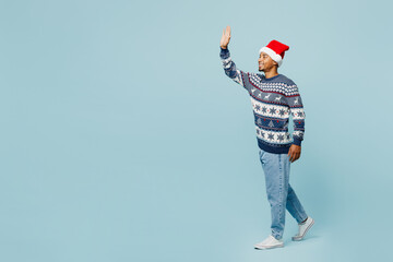 Full body side view young man wear knitted sweater Santa hat posing walk go strolling waving hand isolated on plain pastel blue background. Happy New Year 2024 celebration Christmas holiday concept. - obrazy, fototapety, plakaty
