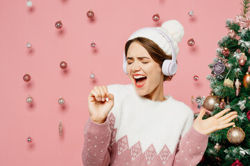 Merry fun young woman wearing white sweater hat posing listening to music in headphones sing song...