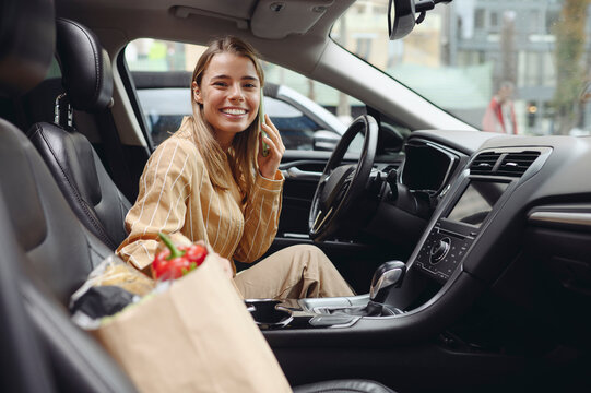 Young fun customer woman wear casual clothes talk mobile cell phone driving car with bag of goods after shopping buying products at supermaket store grocery shop hypermarket. Purchasing food concept.