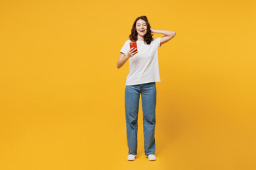 Full body young amazed impressed happy woman she wear white blank t-shirt casual clothes hold head use mobile cell phone isolated on plain yellow orange background studio portrait. Lifestyle concept. - obrazy, fototapety, plakaty