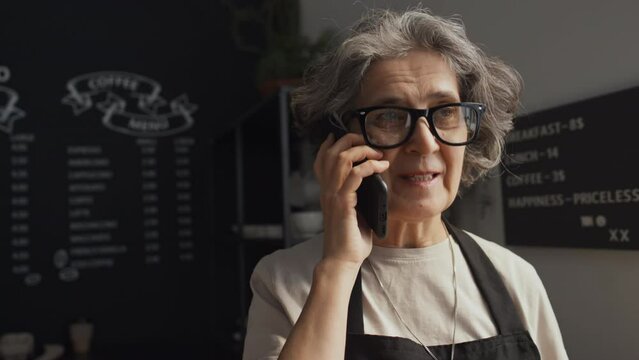 Medium close-up shot of elderly Caucasian woman wearing black apron standing in coffee house talking over phone