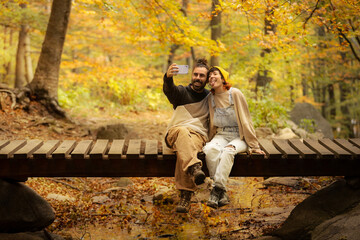 male and female couple talking on the phone, taking selfie in the forest with sitting on a bridge