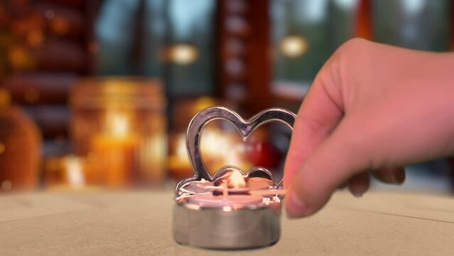 A woman's hand lighting a rose shaped candle, cozy and romantic cabin interior illuminated by the warm glow of candles and heart-shaped balloons, creating a Valentine's Day celebration.