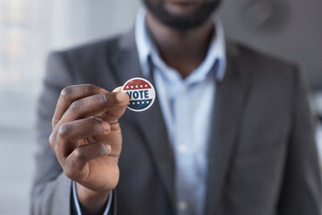 Hand of young African American male citizen of USA showing small round vote badge while standing in...