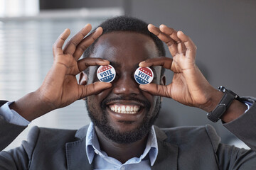 Happy young African American voter with wide toothy smile holding two small round vote badges by...