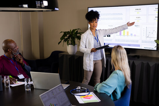 Happy diverse female doctor with tablet giving presentation to colleagues at office meeting