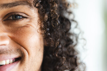Half face of happy biracial man with long dark curly hair smiling in sunny living room, copy space