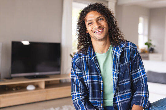 Happy biracial man with long curly hair sitting on sofa and smiling in sunny living room at home - Powered by Adobe