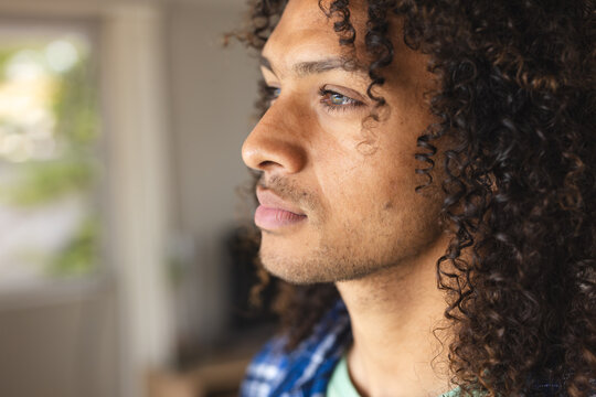 Pensive biracial man with long curly hair looking ahead in sunny living room at home