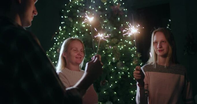 A happy family celebrates the new year at the Christmas tree, holding burning sparklers