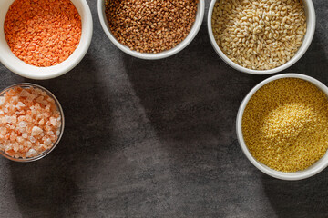 Raw buckwheat, wheat, red lentils and pink salt in bowls, on dark background, top view, space to copy text