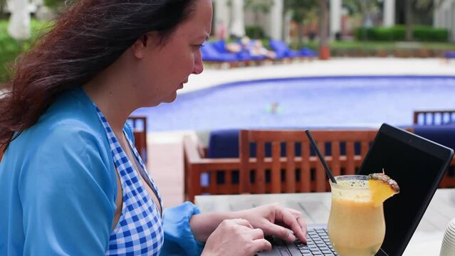 Dedicated woman working on laptop in luxurious hotel outdoor cafe. Female freelancer is having drink while sitting against swimming pool during windy day. She is enjoying summer holiday.